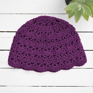 Puede incluir: Un gorro de ganchillo de color morado oscuro con un patr&oacute;n repetitivo de puntos de concha en toda la tela. El gorro tiene una corona redondeada y un borde festoneado en la parte inferior, mostrando un dise&ntilde;o calado y texturizado.
