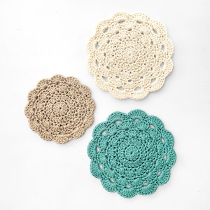 May include: Three handmade crochet coasters in a top-down view. One is beige, one is teal, and the other is tan. Each coaster has a scalloped edge and a spiral center. The coasters are made of yarn.