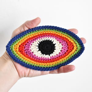 May include: A crocheted eye with a rainbow pattern and a black pupil. The eye is made with colorful yarn and has a white center.