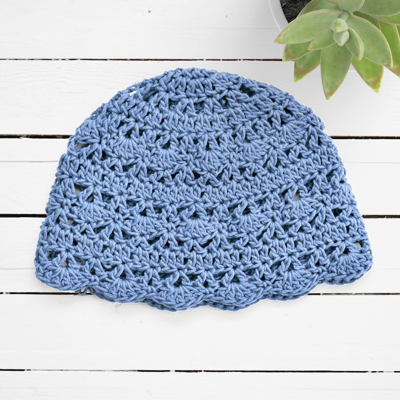 Puede incluir: Un gorro de ganchillo en color azul polvoriento que presenta un patr&oacute;n de puntos en forma de abanico repetido en todo el dise&ntilde;o. La prenda tiene una corona redondeada y un borde festoneado, confeccionado con hilo texturizado en un tono azul s&oacute;lido.