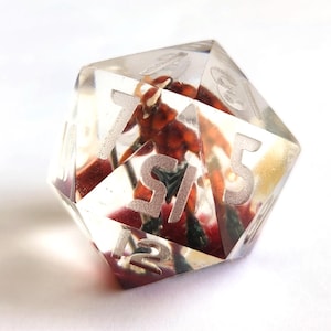 May include: A clear, multi-sided dice with numbers 1 through 6 etched on each side. The dice is filled with red, brown, and green resin, creating a unique and colorful design.