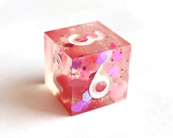 May include: A pink and clear resin dice with white numbers 3 and 6. The dice is filled with pink and purple glitter and hearts.