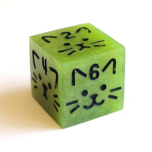 May include: A green dice with black cat faces on each side. The dice is made of a translucent material, and the cat faces are visible through the material.