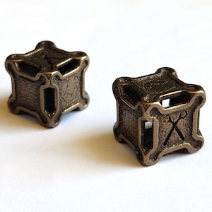 May include: Two bronze-colored, cube-shaped beads with a textured surface. Each bead features a cross design and a rectangular opening. The beads are likely for jewelry making or crafts.