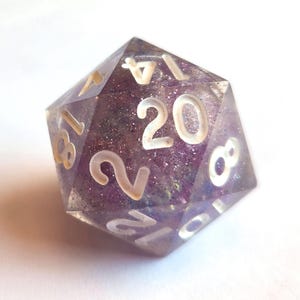 May include: A close-up of a twenty-sided die, or D20, with a purple and glittery interior. The numbers are white and clearly visible. This gaming accessory is perfect for tabletop role-playing games.