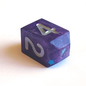 May include: A purple resin die with silver numbering. The die features the number 4 on top and the number 2 on the side. A small blue star is visible on one side. The die is designed for tabletop games.