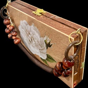 May include: A wooden clutch with a large white rose painted on the front. The rectangular box-shaped purse has a brown leather strap and wooden bead accents. A gold-colored clasp and hardware complete the design.
