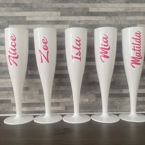 May include: Five white plastic champagne flutes with pink lettering. The names Alice, Zoe, Isla, Mia, and Matilda are written on the flutes.