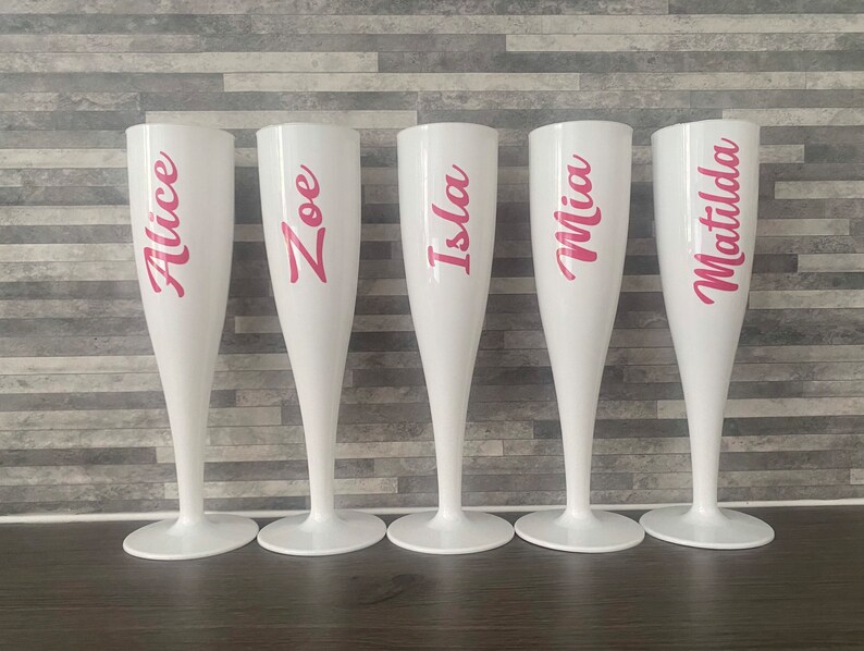 May include: Five white plastic champagne flutes with pink lettering. The names Alice, Zoe, Isla, Mia, and Matilda are written on the flutes.
