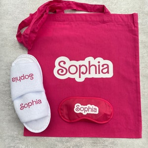 May include: A pink tote bag with the name "Sophia" printed in white. The bag contains a pair of white slippers with the name "Sophia" printed in pink and a pink sleep mask with the name "Sophia" printed in white.