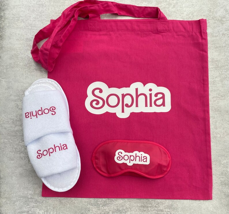 May include: A pink tote bag with the name "Sophia" printed in white. The bag contains a pair of white slippers with the name "Sophia" printed in pink and a pink sleep mask with the name "Sophia" printed in white.