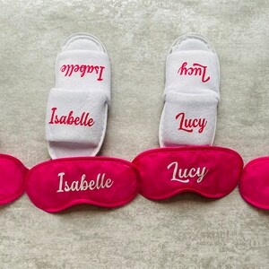 May include: Four pairs of white slippers with pink eye masks. Each pair of slippers and eye mask has a name embroidered on them: Elsie, Isabelle, Lucy, and Eva.