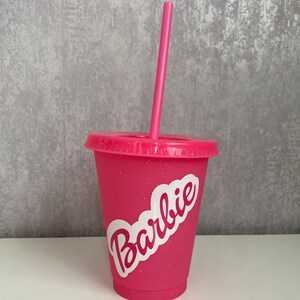 May include: Pink plastic tumbler with a white glitter finish and a white sticker that says "Barbie". The tumbler has a pink straw.
