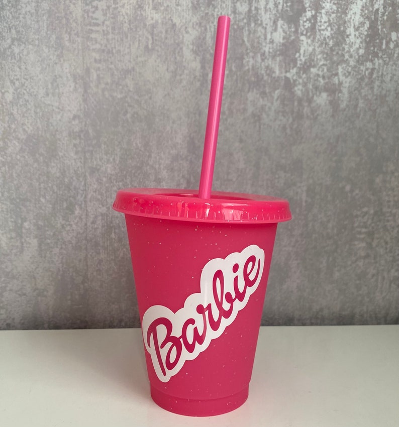 May include: Pink plastic tumbler with a white glitter finish and a white sticker that says "Barbie". The tumbler has a pink straw.