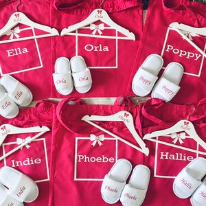 May include: Six red canvas tote bags with white handles and white personalized text. Each bag has a white wooden hanger with a pink bow and the name of the person the bag is for. The names are Ella, Orla, Poppy, Indie, Phoebe, and Hallie. Each bag also has a pair of white slippers with the name of the person the bag is for.