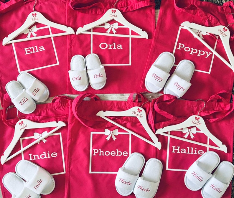 May include: Six red canvas tote bags with white handles and white personalized text. Each bag has a white wooden hanger with a pink bow and the name of the person the bag is for. The names are Ella, Orla, Poppy, Indie, Phoebe, and Hallie. Each bag also has a pair of white slippers with the name of the person the bag is for.