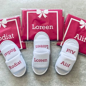 May include: Three pairs of white slippers with pink embroidered names: Rodiat, Loreen, and Alif. Each pair is packaged in a pink satin bag with a white bow and the name embroidered on the front.