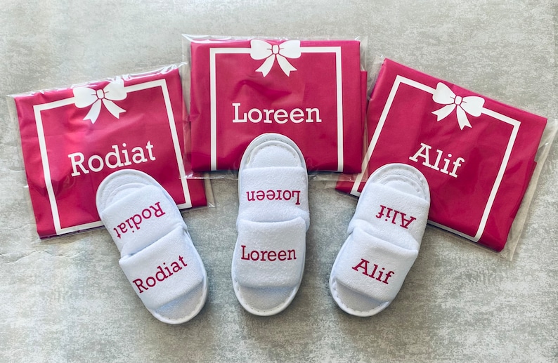 May include: Three pairs of white slippers with pink embroidered names: Rodiat, Loreen, and Alif. Each pair is packaged in a pink satin bag with a white bow and the name embroidered on the front.