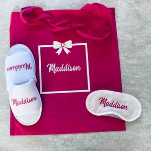 May include: A pink tote bag with a white box that says "Maddison" and a bow. The bag also includes a pair of white slippers with the name "Maddison" and a white sleep mask with the name "Maddison".