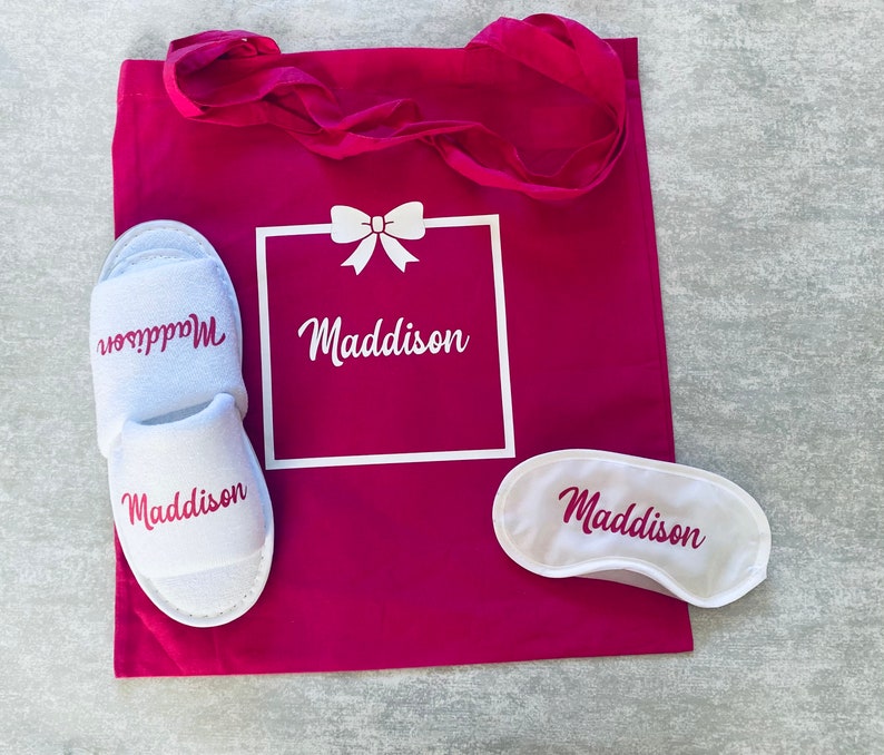 May include: A pink tote bag with a white box that says "Maddison" and a bow. The bag also includes a pair of white slippers with the name "Maddison" and a white sleep mask with the name "Maddison".