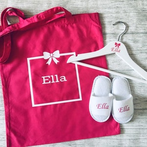 May include: A pink canvas tote bag with white handles and the name "Ella" printed on it. The bag is next to a white wooden hanger with the name "Ella" printed on it. There are also two white slippers with the name "Ella" embroidered on them.
