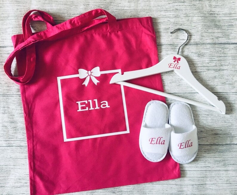May include: A pink canvas tote bag with white handles and the name "Ella" printed on it. The bag is next to a white wooden hanger with the name "Ella" printed on it. There are also two white slippers with the name "Ella" embroidered on them.