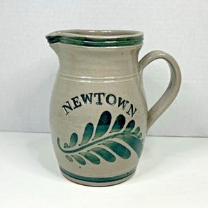 May include: A beige ceramic pitcher featuring a dark green painted fern leaf design and the word NEWTOWN in dark green lettering. The pitcher has a curved handle, a pouring spout, and dark green bands around the rim and the base of the vessel.