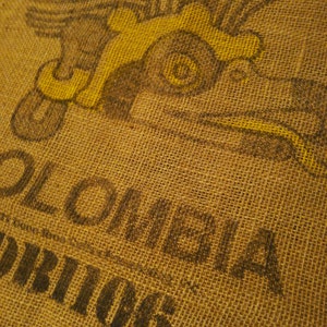 May include: A brown burlap sack with a yellow and black printed design of a flying bird. The text "Colombia" and "Orions" is printed on the sack.
