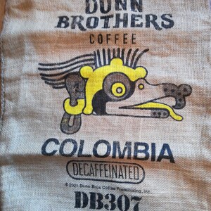 May include: A burlap coffee bag with the text "Dunn Brothers Coffee" and a yellow and brown graphic of a stylized bird. The bag also says "Colombia Decaffeinated" and "DB307".