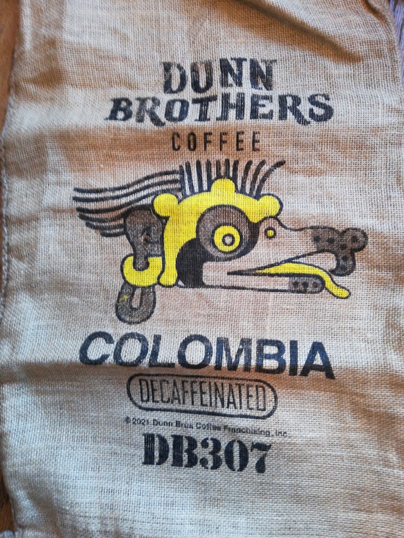 May include: A burlap coffee bag with the text "Dunn Brothers Coffee" and a yellow and brown graphic of a stylized bird. The bag also says "Colombia Decaffeinated" and "DB307".
