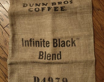 May include: A rectangular tan burlap coffee sack featuring black printed text that reads DUNN BROS COFFEE, Infinite Black Blend, and the alphanumeric code D4979. The woven fabric has a coarse, natural texture with visible fibers throughout the surface.
