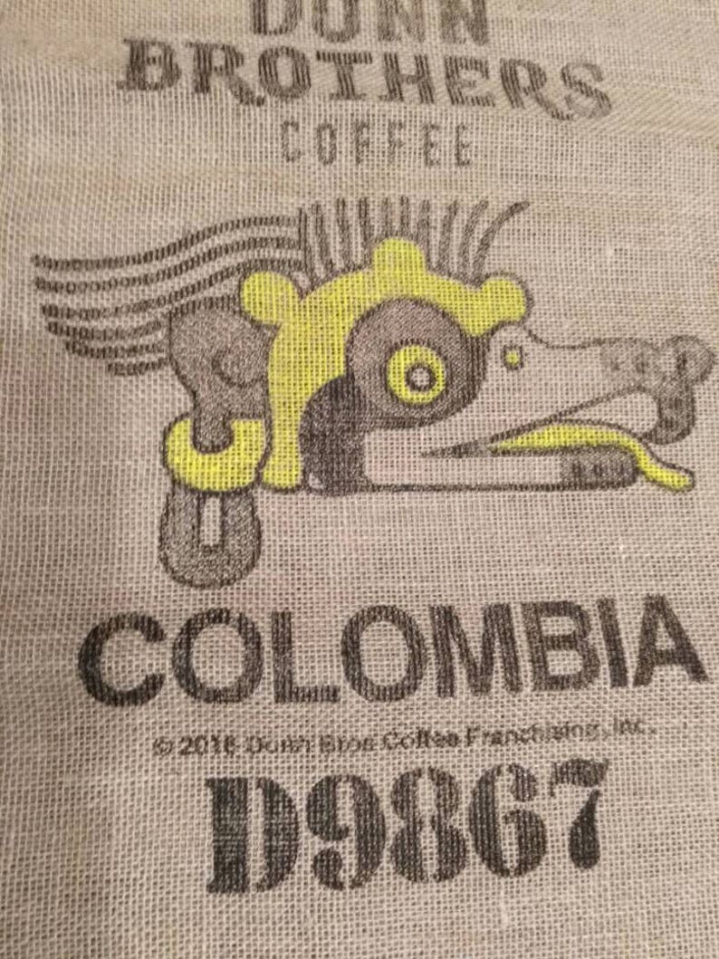 May include: A brown burlap coffee bag with the text "Dunn Brothers Coffee" and a stylized image of a bird with yellow accents. The bag also has the text "Colombia" and the year "1986".