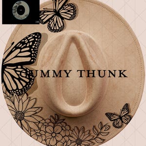 May include: Tan fedora hat with black butterfly and floral designs. The hat features the text "UMMY THUNK" in black. The hat has a wide brim and a pinched crown. The hat is decorated with butterflies and flowers.