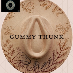 May include: A beige fedora hat featuring brown illustrations of bees, leaves, and a mushroom. The text "GUMMY THUNK" is printed in black across the hat. A small image of a circular design is in the upper left corner.