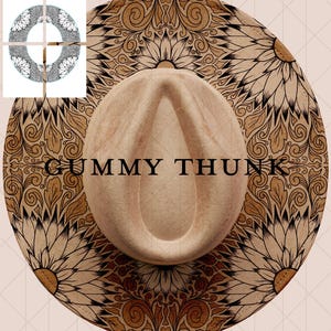 Op de afbeelding: Beige fedora hoed met een brede rand met een bloemen- en wervelpatroon in bruin- en beigetinten. De tekst "GUMMY THUNK" is op de rand gedrukt. De hoed heeft een klassieke druppelvormige kroon.