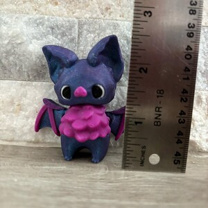 Pode incluir: Uma pequena figura de morcego roxo e azul com pele rosa ao redor do pesco&ccedil;o. A figura tem aproximadamente 5 cm de altura e est&aacute; em p&eacute; sobre uma superf&iacute;cie de madeira.