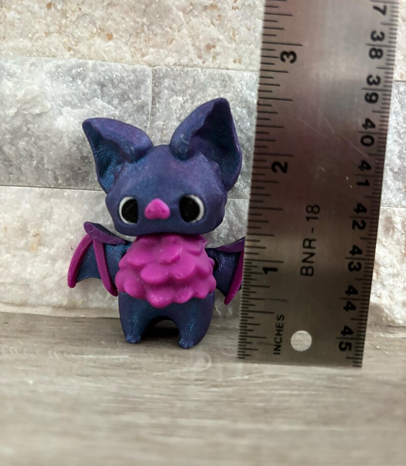Pode incluir: Uma pequena figura de morcego roxo e azul com pele rosa ao redor do pesco&ccedil;o. A figura tem aproximadamente 5 cm de altura e est&aacute; em p&eacute; sobre uma superf&iacute;cie de madeira.