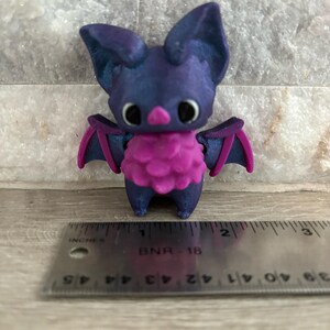 Pode incluir: Uma pequena figura de morcego impressa em 3D de cor roxa e rosa. O morcego tem orelhas grandes, um corpo peludo rosa e olhos pretos.