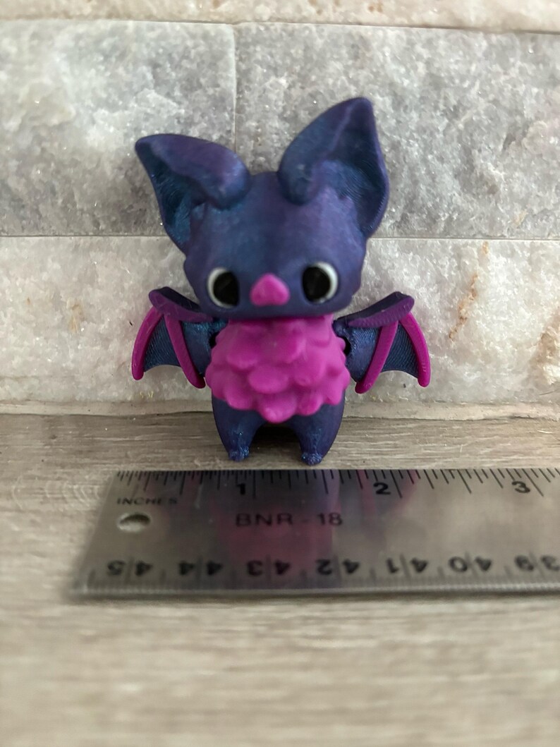 Pode incluir: Uma pequena figura de morcego impressa em 3D de cor roxa e rosa. O morcego tem orelhas grandes, um corpo peludo rosa e olhos pretos.
