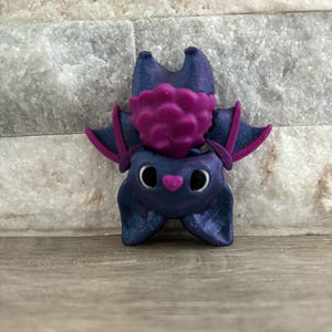 Pode incluir: Uma pequena figura de morcego roxo e preto com uma cabe&ccedil;a texturizada rosa e roxa. O morcego tem grandes olhos pretos e um nariz rosa.