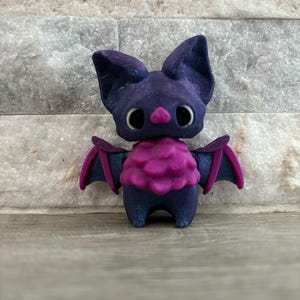 Pode incluir: Uma pequena figura de morcego impressa em 3D de cor roxa e rosa com olhos grandes e um peito rosa felpudo.