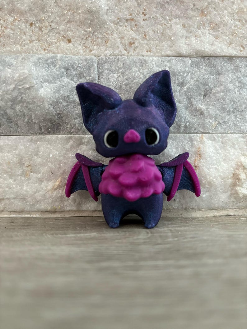Pode incluir: Uma pequena figura de morcego impressa em 3D de cor roxa e rosa com olhos grandes e um peito rosa felpudo.