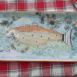 Peut inclure: Plat à servir rectangulaire en céramique avec un motif de poisson. Le poisson est orange et jaune avec des taches noires, sur un fond bleu et vert. Le plat a un bord marron et est décoré d'épis de blé.