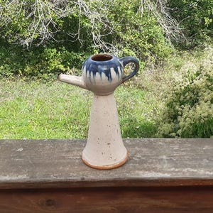 Pode incluir: Uma chaleira de cerâmica artesanal com um design único. A chaleira tem uma base creme e um topo esmaltado em azul escuro e branco. Possui um bico e uma alça, em um cenário natural ao ar livre.