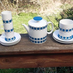Op de afbeelding: Een witte theepot en drie witte kopjes met blauwe en gouden geometrische patronen. De theepot heeft een blauw deksel. De kopjes zijn op elkaar gestapeld. De kopjes en de theepot staan op een houten oppervlak.