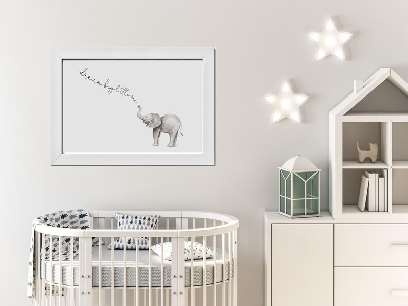 May include: A white framed print with a watercolor illustration of a grey elephant with the text "dream big little one" above it. The print is hanging on a white wall with two white star-shaped lights above it.