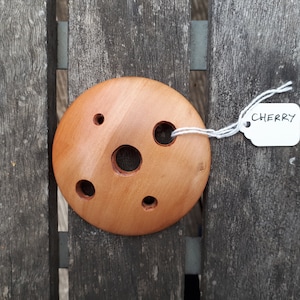 Peut inclure: Un disque rond en bois avec cinq trous découpés. Le disque est de couleur brun clair et a une finition lisse et polie. Le disque est étiqueté "CHERRY".