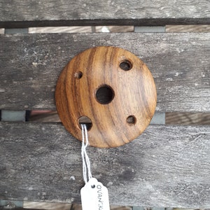 May include: A round, wooden object with a central hole and four smaller holes around the perimeter. The wood grain is visible, and a white tag with text is attached. The object is likely a decorative or functional item.
