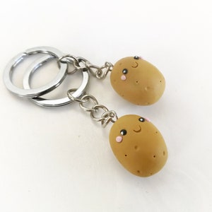 Op de afbeelding: Een zilveren sleutelhanger met twee bruine aardappelcharms. Elke aardappel heeft een schattig cartoon gezicht met zwarte ogen, een roze blos en een glimlachende mond.