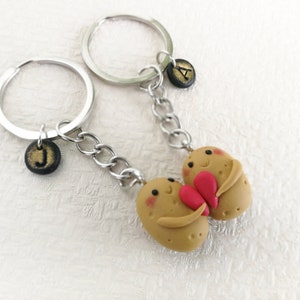 以下が含まれることがあります： 顔と腕のあるポテトの形をしたキーホルダー2つが、赤いハートを持っています。キーホルダーは、黒い文字のチャームが付いたシルバーのキーリングに取り付けられています。